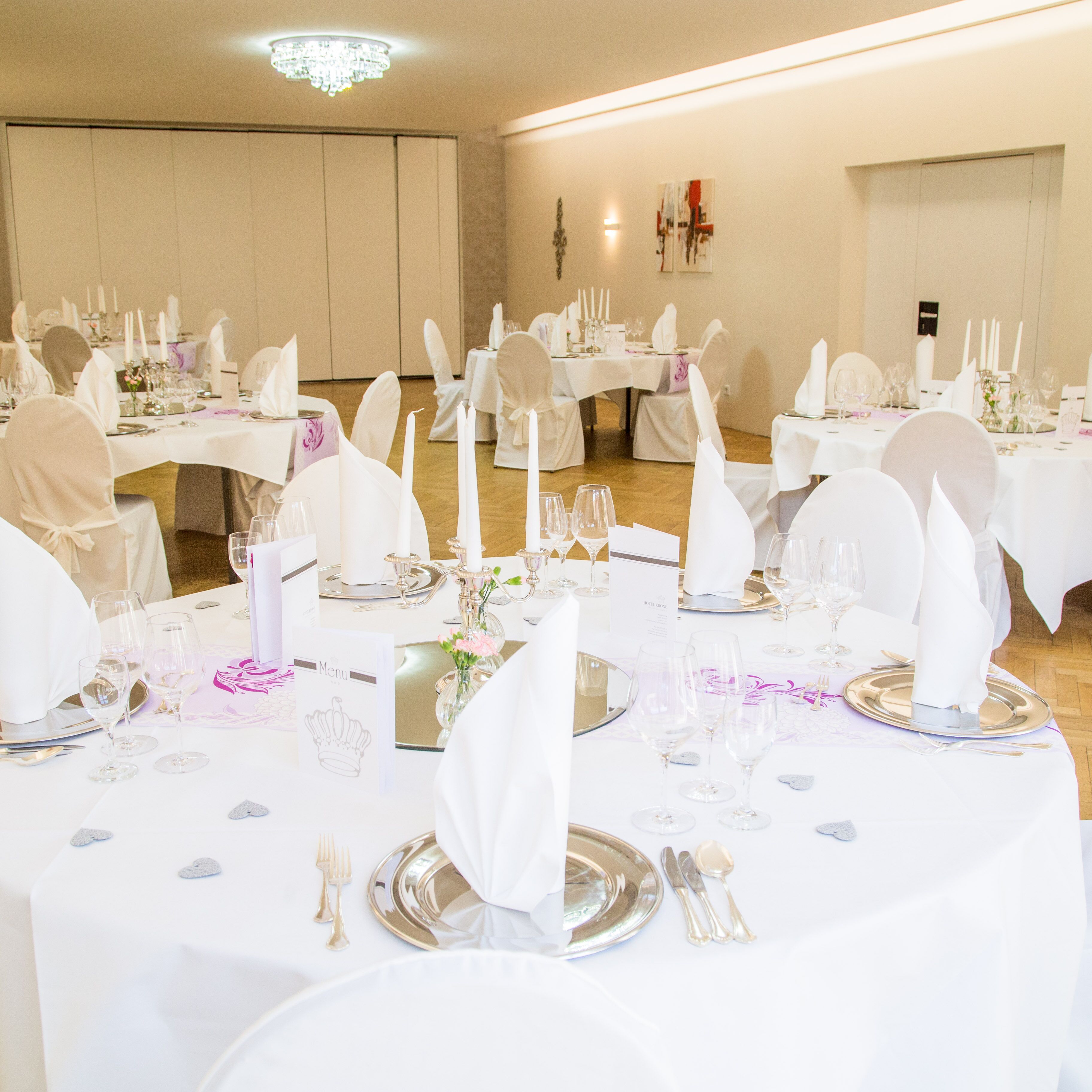 Une salle de banquet avec des tables rondes dressées pour un événement formel, agrémentée de nappes blanches, de couverts en argent et de verres.