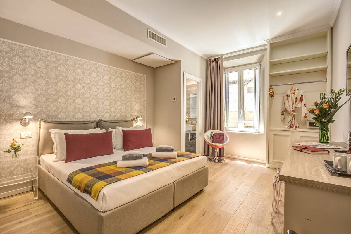 A well-lit bedroom with a large bed, a bedside table, a mirror, and a window with curtains.