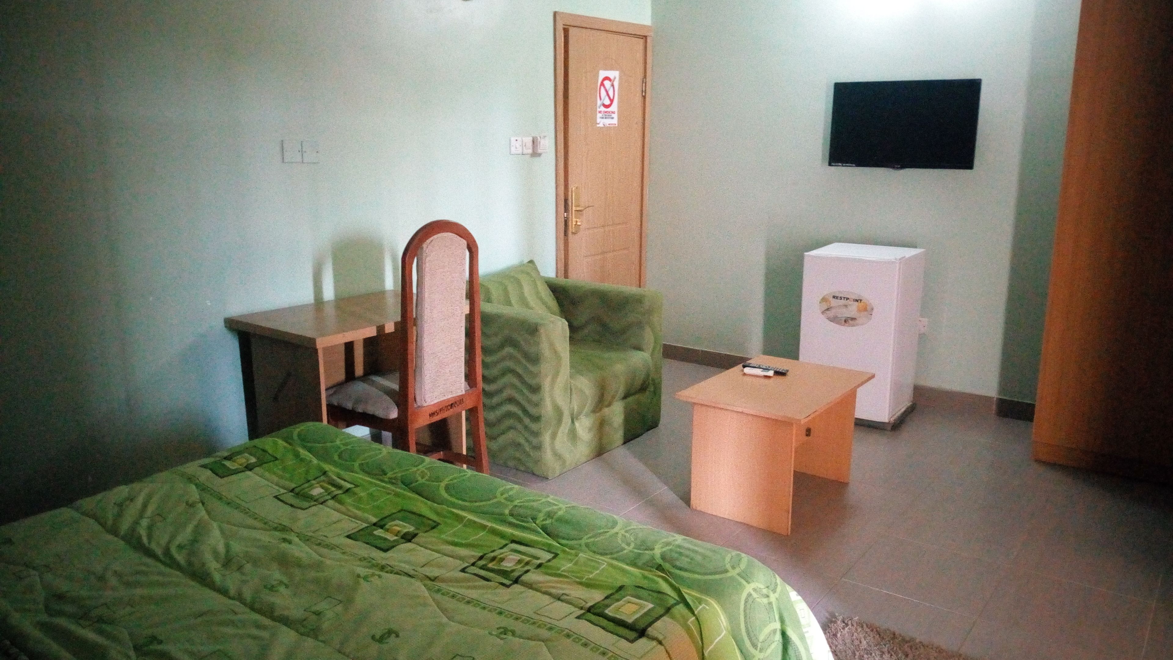 Una habitación pequeña y sencilla con una cama, una silla, una mesita, televisión y un refrigerador.