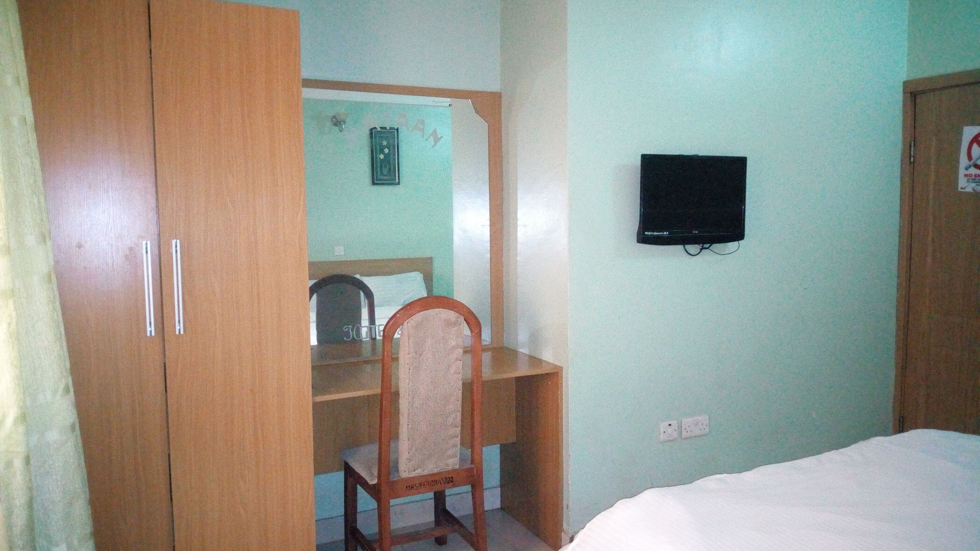 A bedroom with a bed, a wooden wardrobe, a desk with a chair, a television on the wall, and a light switch.