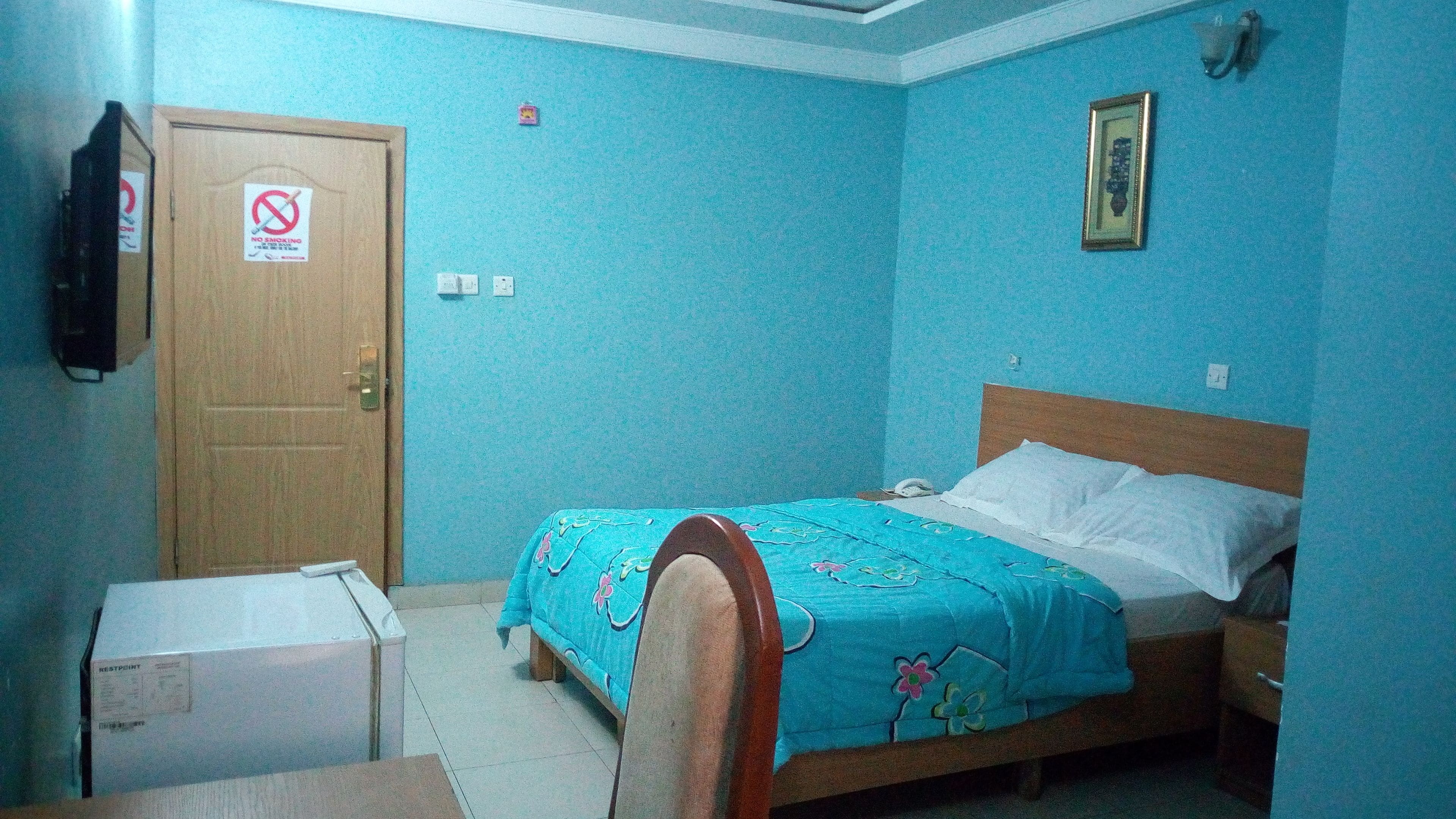 A bedroom with a bed, a wooden door, a small table, and a wall-mounted light fixture.