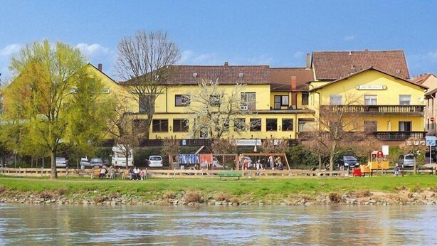 Un bâtiment jaune avec un toit en tuiles, une enseigne indiquant « Gasthof », et une rivière au premier plan.