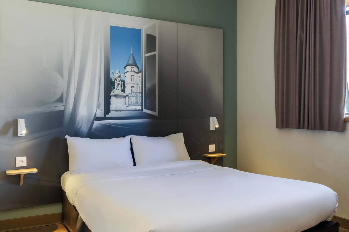 A hotel room with a large bed, two bedside tables with lamps, a wall-mounted TV, and a painting of a building.