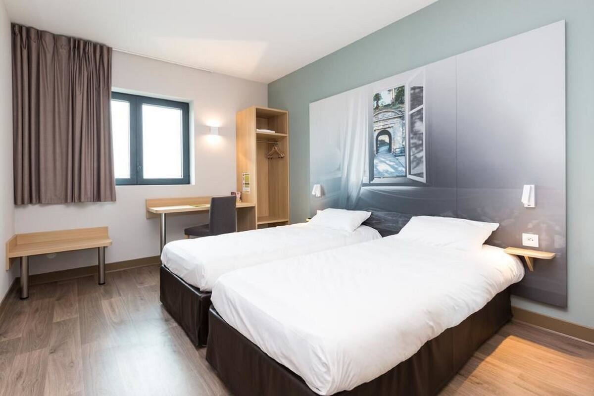 A modern hotel room with two beds, a desk, a chair, a wardrobe, and a large window.