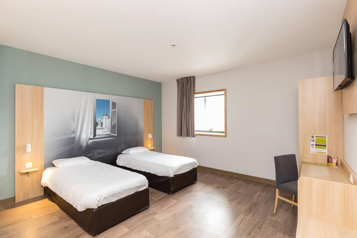 A hotel room with two beds, a desk, a chair, a TV, and a window with curtains.