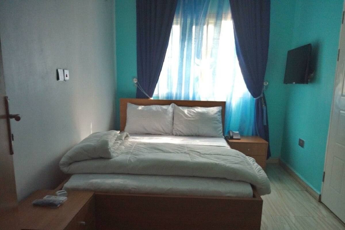 Un dormitorio con cama, mesitas de noche, un banco de madera, televisor en la pared y una ventana con cortinas.