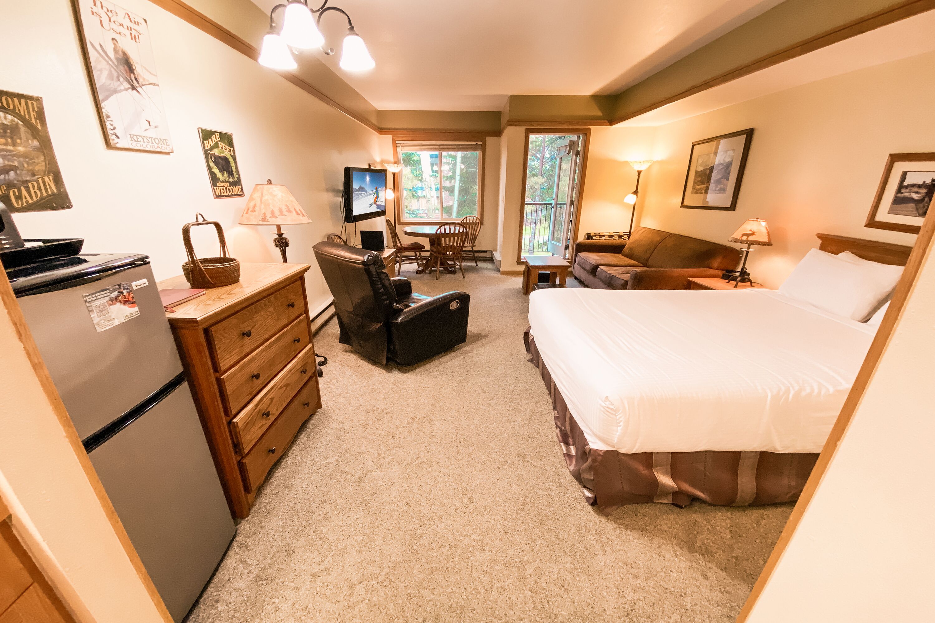 A compact hotel room with a bed, a desk, a chair, a television, and a refrigerator.