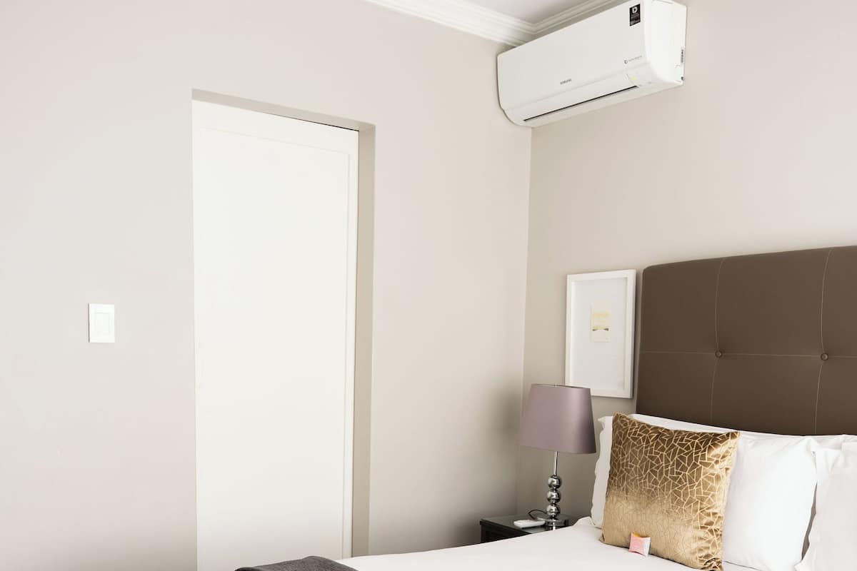 A hotel room with a bed, bedside table, lamp, and an air conditioning unit.
