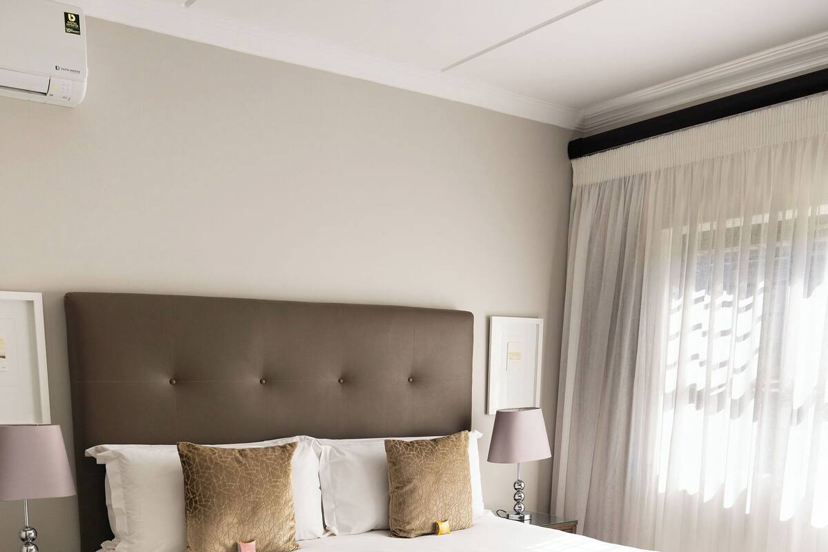 A hotel room with a large bed, two bedside tables, a wall-mounted air conditioner, and a window with sheer curtains.