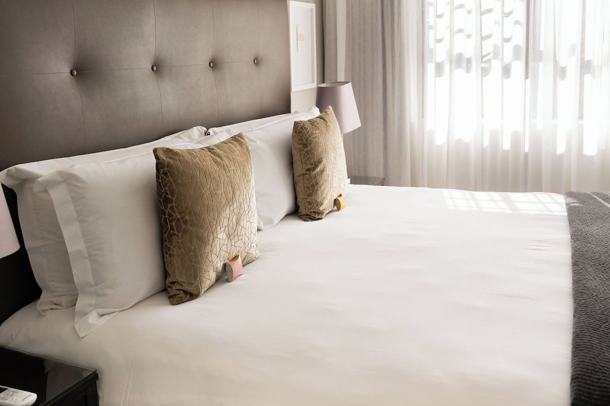 A neatly made bed with white linens and two decorative pillows, a dark headboard, a bedside table with a lamp, and sheer curtains allowing natural light.