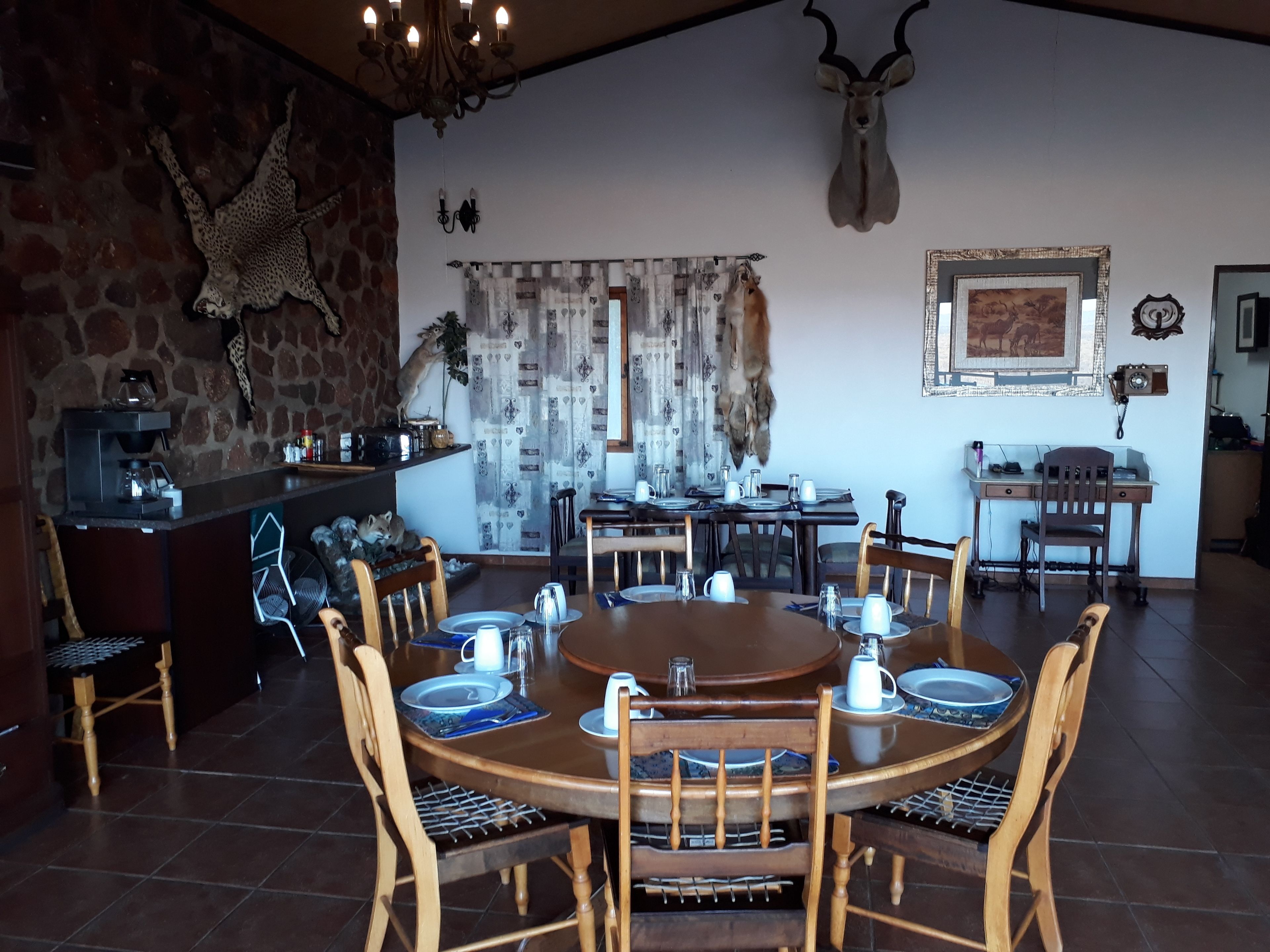 A dining area with a round wooden table set for four, surrounded by wooden chairs. A stone wall and a chandelier are visible in the background.