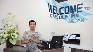 Un mostrador de recepción con un cartel que dice "WELCOME TO CAPULE INN MEDAN", una impresora, un monitor de computadora que muestra imágenes de habitaciones y un jarrón con flores blancas.