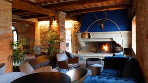 A rustic interior with exposed brick walls, a stone fireplace, and wooden beams. There are leather sofas, a round table, and potted plants.
