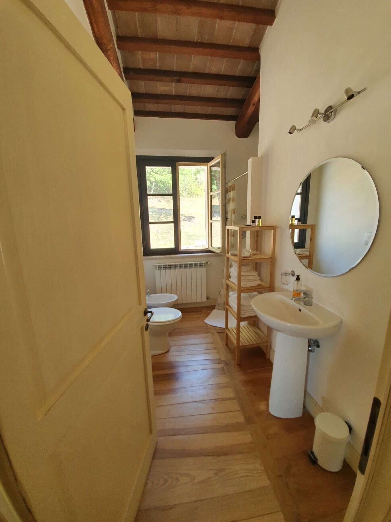 A narrow hallway leading to a bathroom with a wooden floor, a white sink, a mirror, and a window providing natural light.