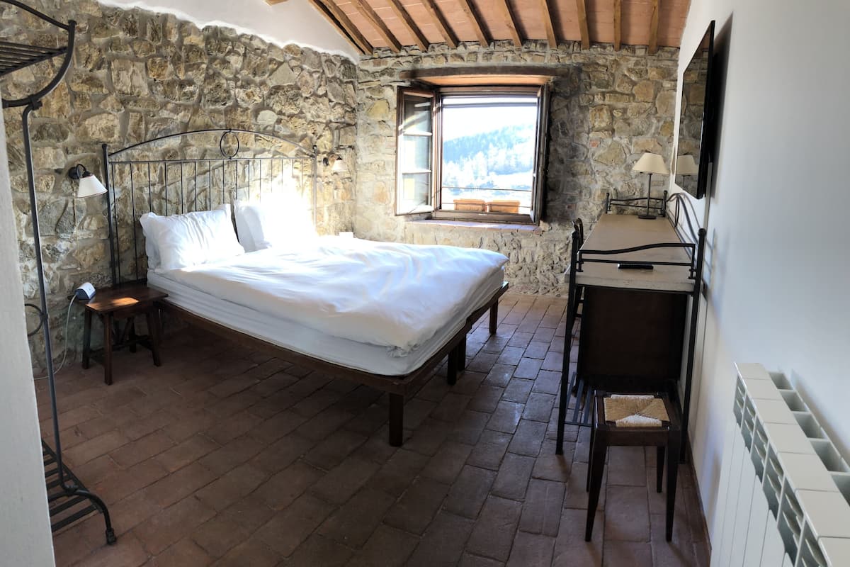 A bedroom with a stone wall, a wooden bed, a small desk with a chair, and a window with a view.