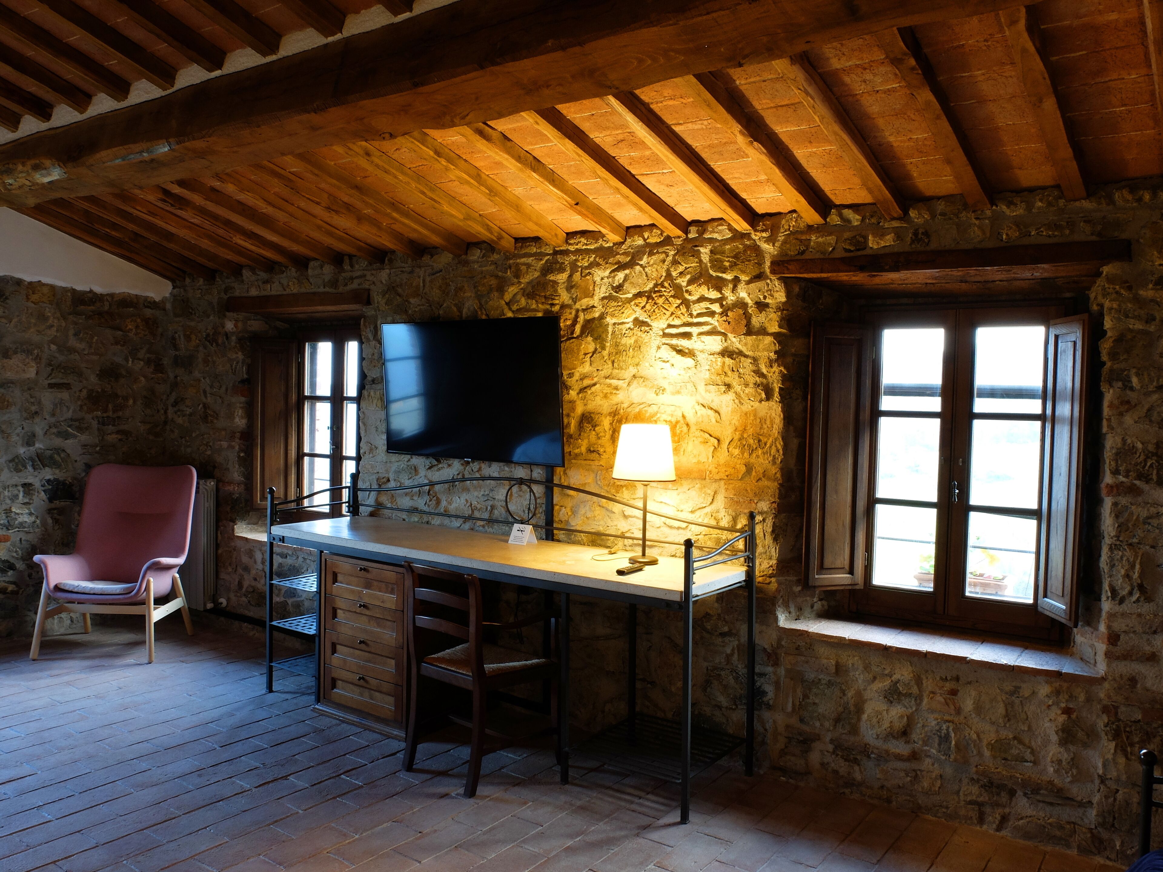 Une pièce rustique aux murs de pierre, avec un plafond en bois, un téléviseur à écran plat fixé au mur, une table avec des chaises, un fauteuil rose et une fenêtre donnant sur un paysage agréable.