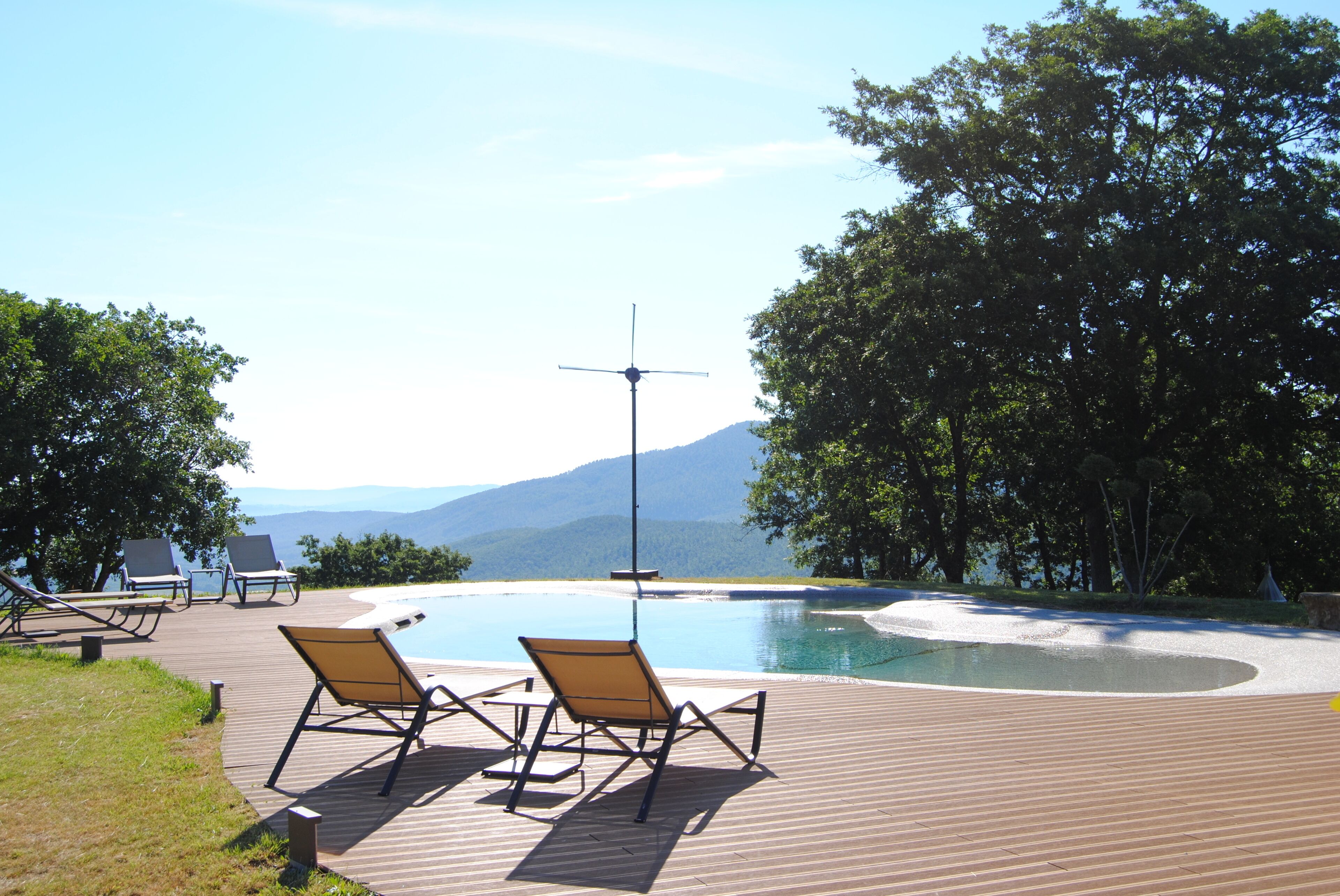 Une terrasse en bois avec des chaises longues et une piscine, entourée d’arbres et avec des montagnes en arrière-plan.