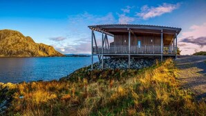 Une cabane en bois dotée d’un balcon donnant sur un lac paisible et sur une formation rocheuse imposante qui se profile à l’horizon.