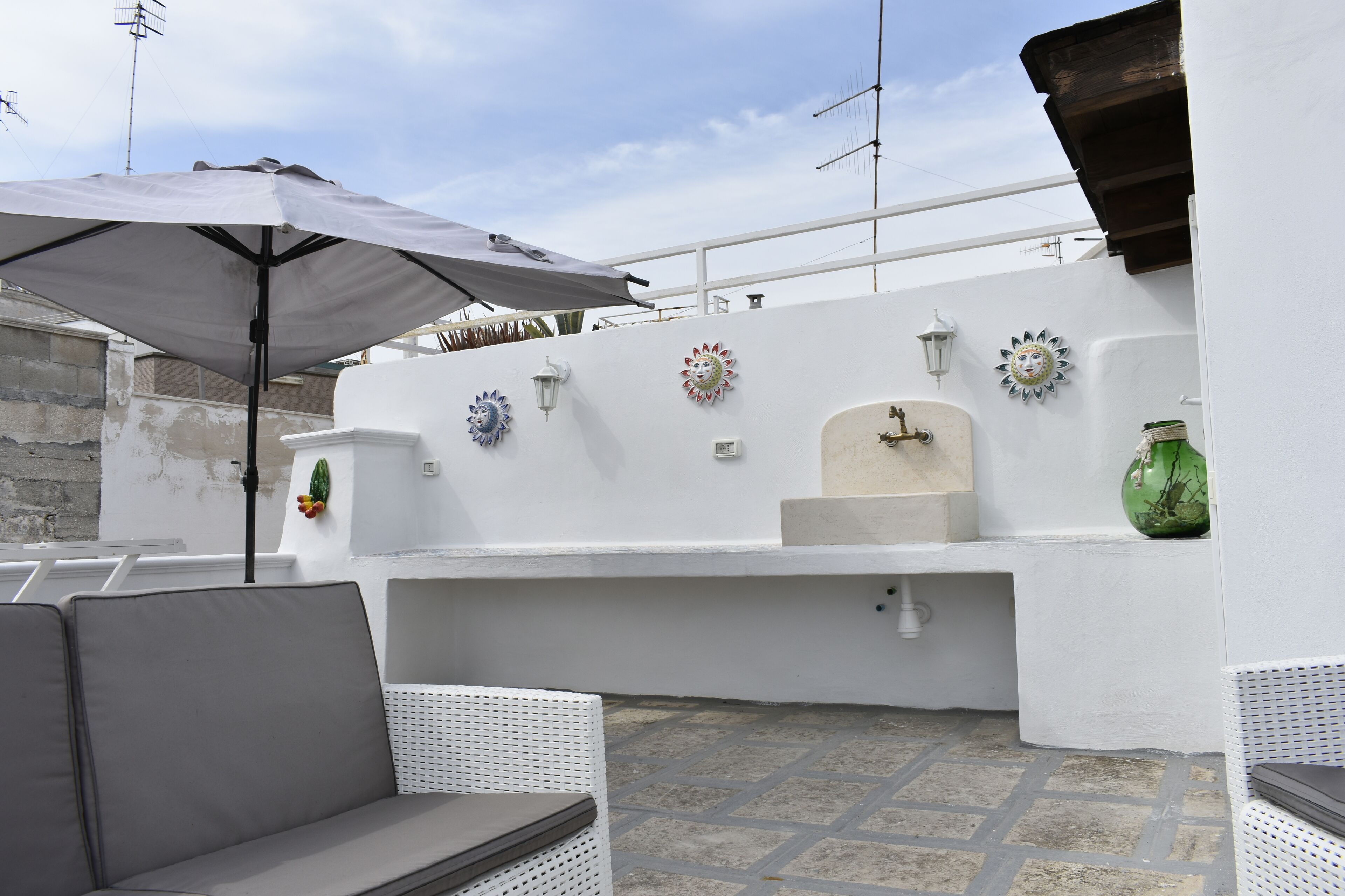 Un espace patio avec un mur blanc, des éléments décoratifs muraux, un évier et un coin salon comprenant une chaise blanche et un parasol.