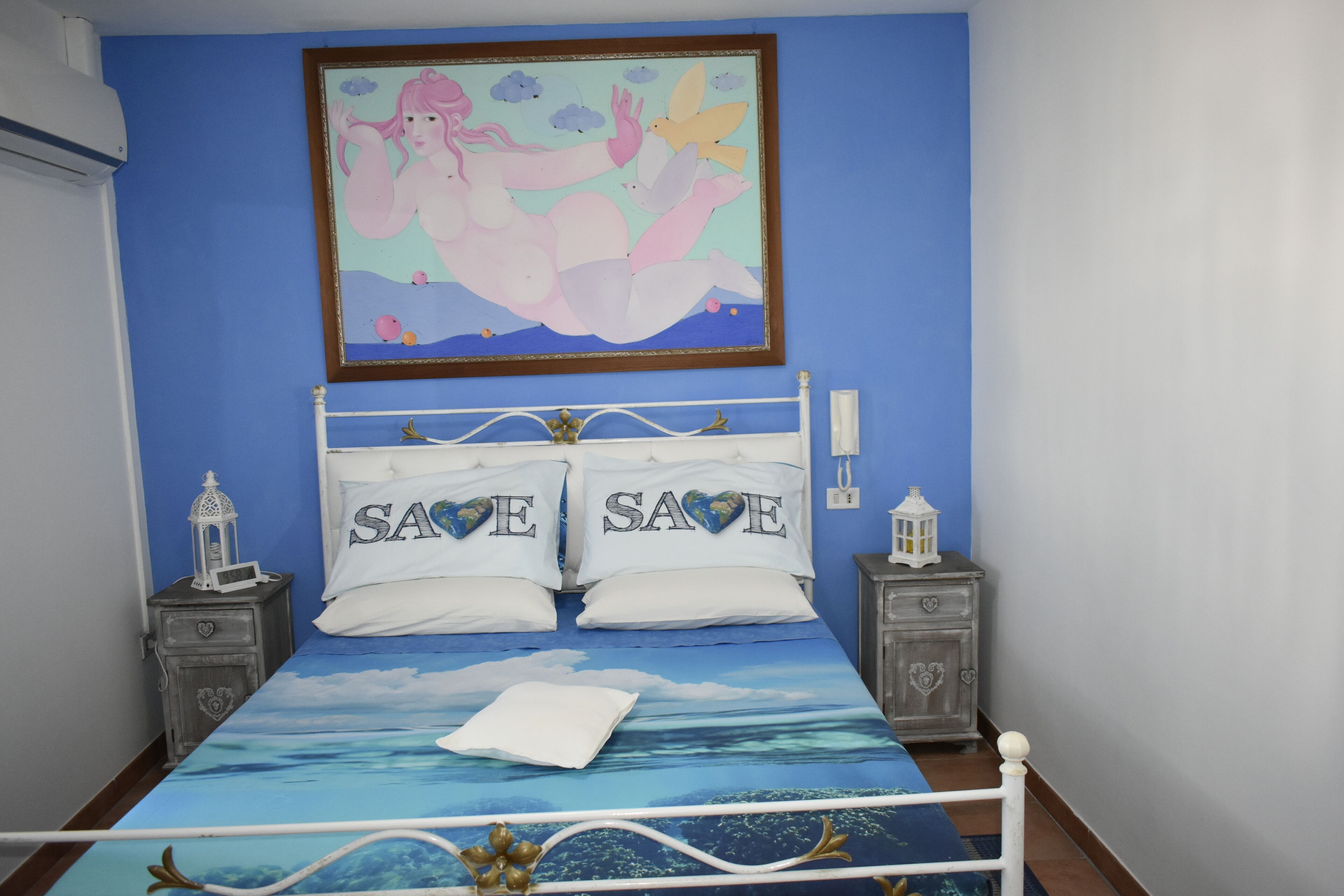 A bedroom with a bed featuring a 'SAVE SAVE' pillow set, a framed artwork above the bed, and a nightstand with a lantern.