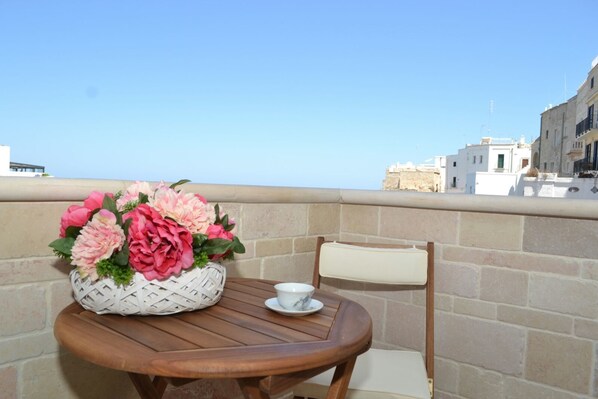 A round wooden table with a white bowl and a woven basket of flowers. A cushioned chair is placed beside the table. The setting is on a balcony with a view of buildings and clear sky. - B&B Prestige (Polignano a Mare)