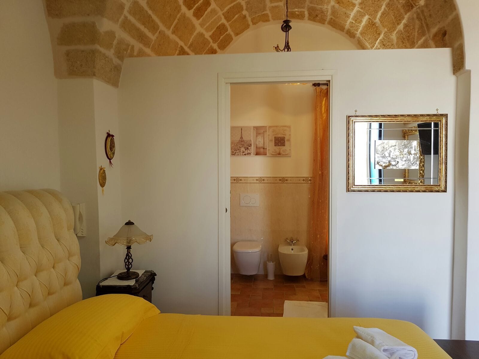 A bedroom with a bed, a nightstand, a lamp, a mirror, and a bathroom visible through an open door.