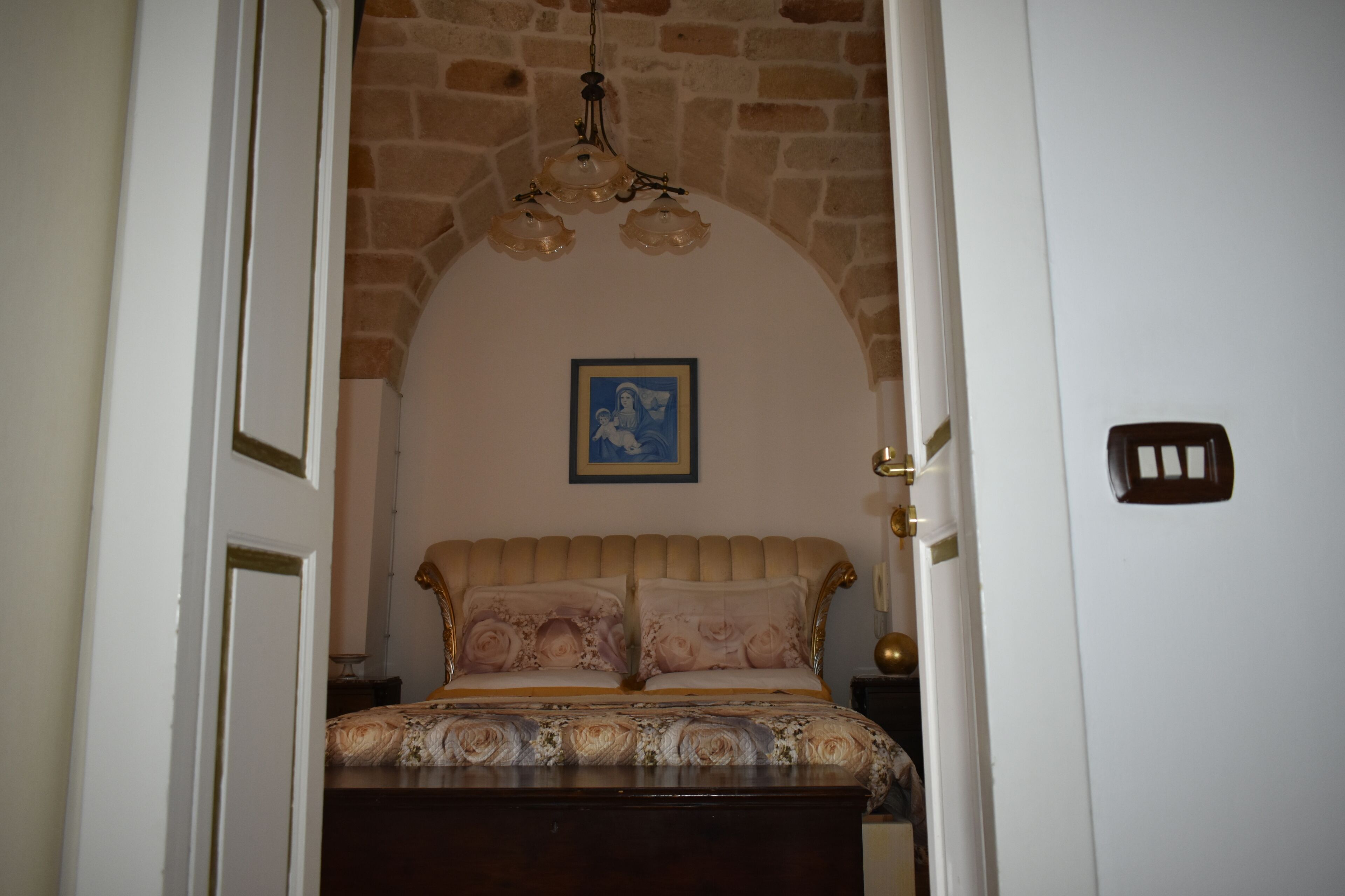 Un dormitorio con cama, una araña, un cuadro enmarcado en la pared y una puerta con interruptor de luz.