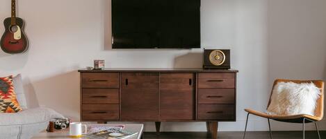 Una sala de estar con una televisión de pantalla plana, una guitarra, un mueble de madera, una mesa de centro con revistas y un sillón con un cojín mullido.