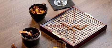 A round wooden table with a chessboard, a glass of amber liquid, a bowl of snacks, and a small dish with a lemon slice.