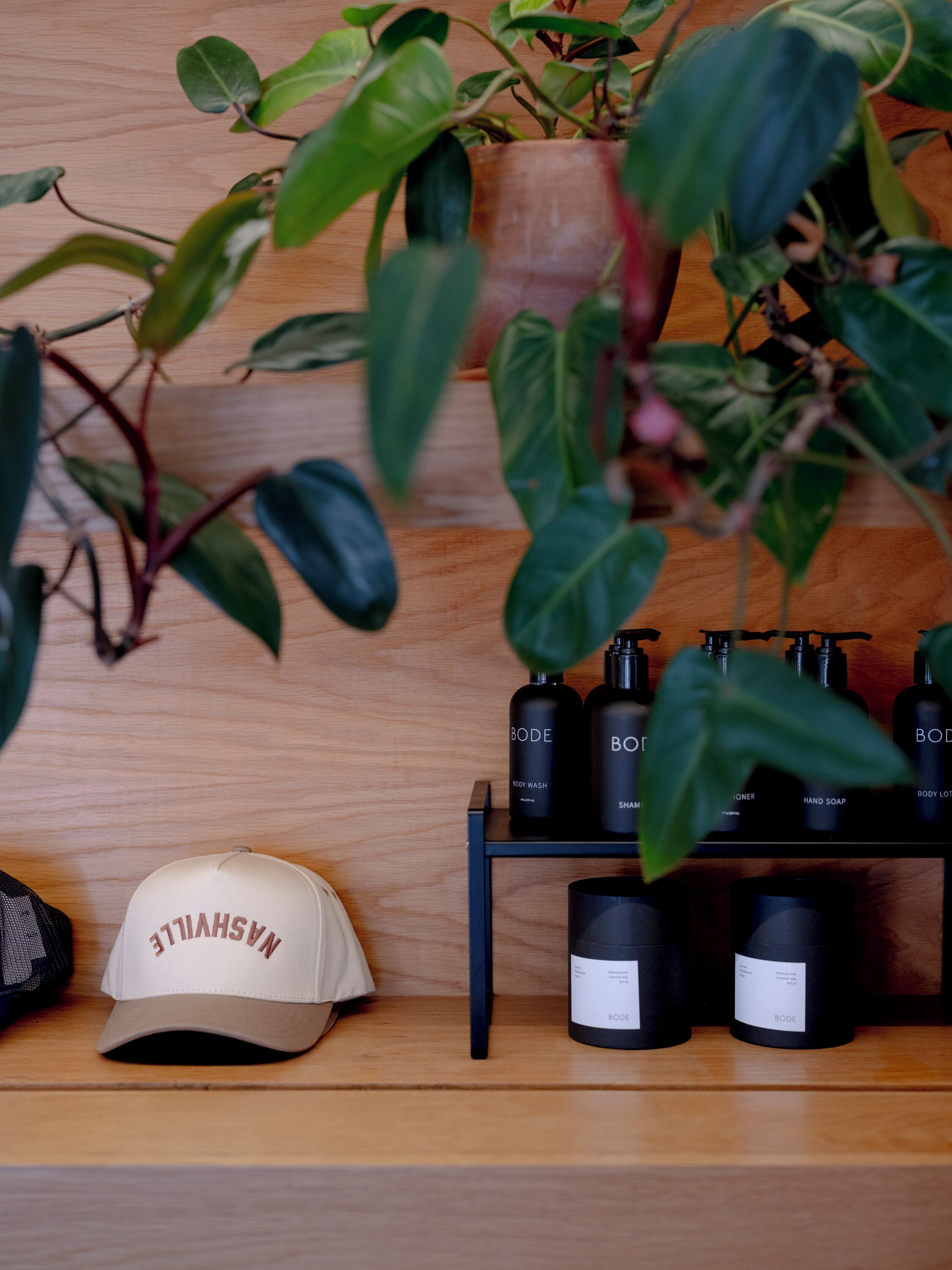 Un estante de madera con una planta, un sombrero y productos de cuidado corporal BODE.