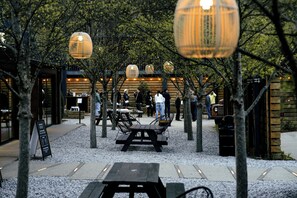 Un espacio al aire libre con mesas y sillas, rodeado de árboles e iluminado por luces colgantes.