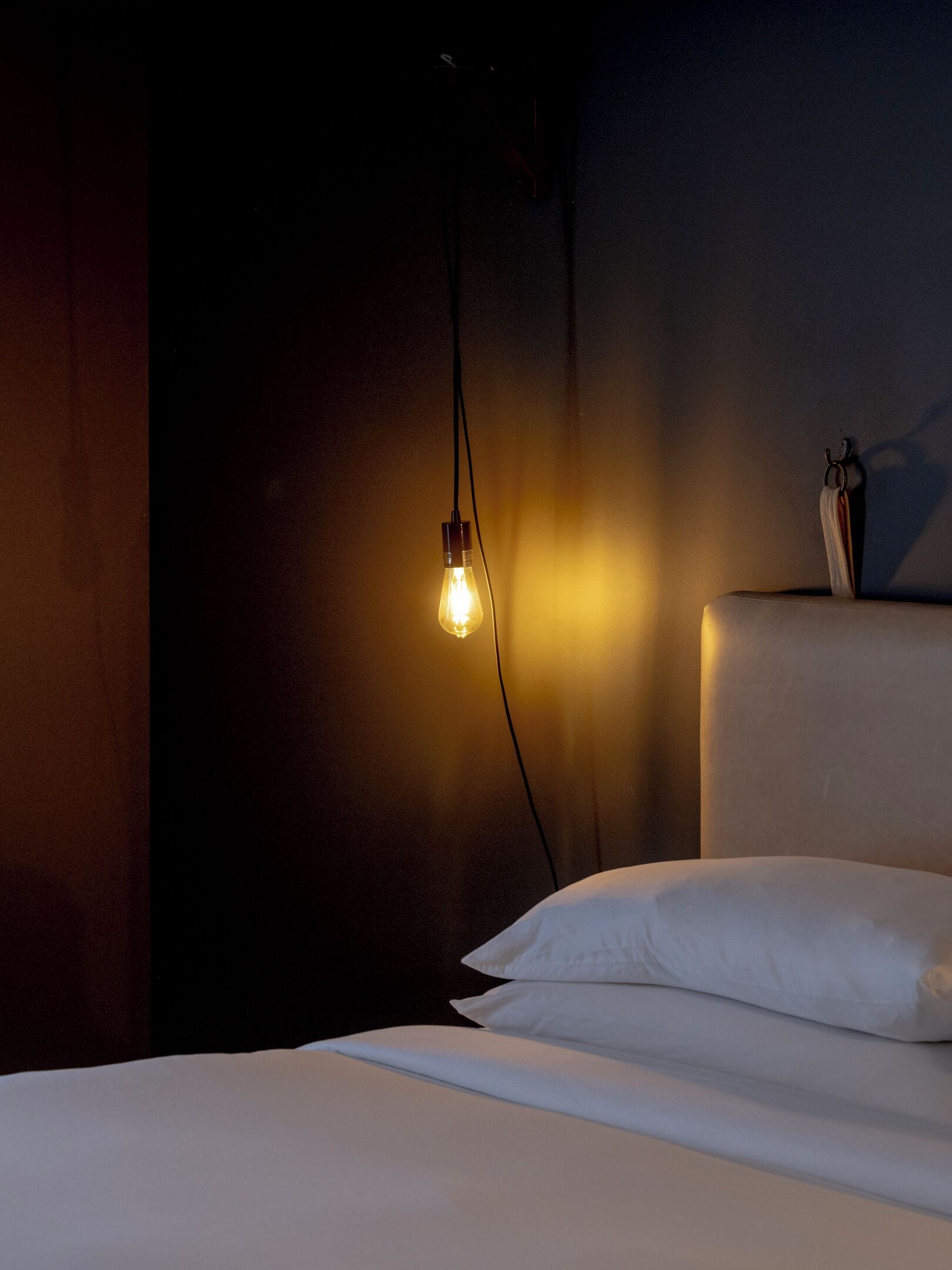 Een schemerig vertrek met een enkele hangende gloeilamp, een wit bed met kussens en een donkere muur.