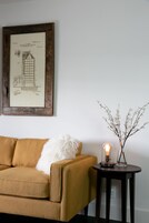 Una habitación con un sofá marrón, un dibujo arquitectónico enmarcado, una mesita con una lámpara y un jarrón con ramas.