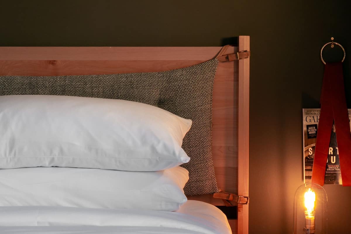 Una cama bien tendida con sábanas blancas, un cabecero de madera y una mesita de noche con una lámpara de estilo vintage.