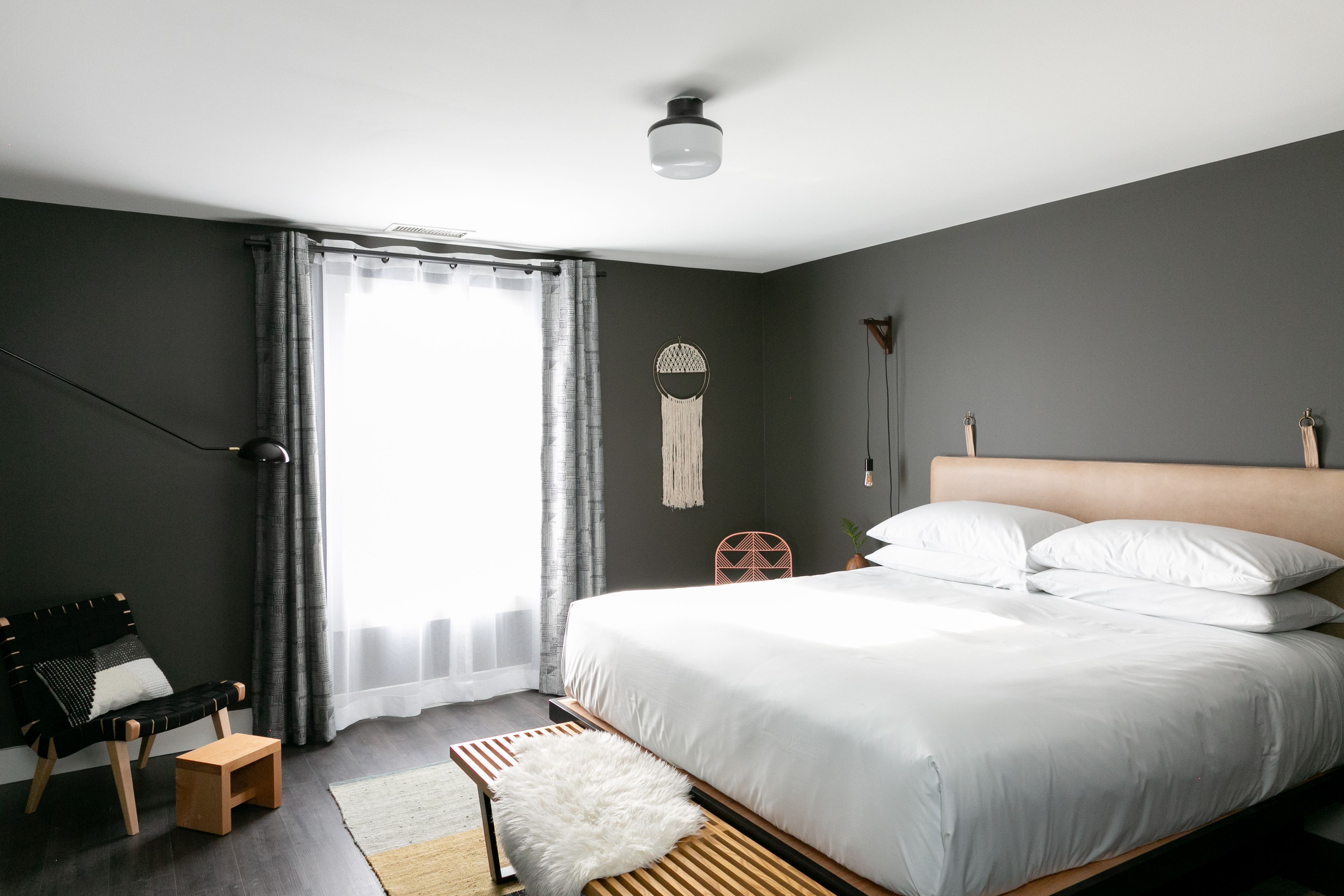 A modern bedroom with a large bed, a small wooden stool, a bench, a macrame wall hanging, and a view of the outdoors through a window with curtains.