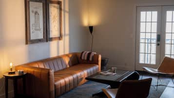 Uma sala de estar moderna com sofá de couro, mesa de centro, mesa lateral com abajur e uma poltrona. Há desenhos arquitetônicos emoldurados na parede, uma janela grande e uma porta.