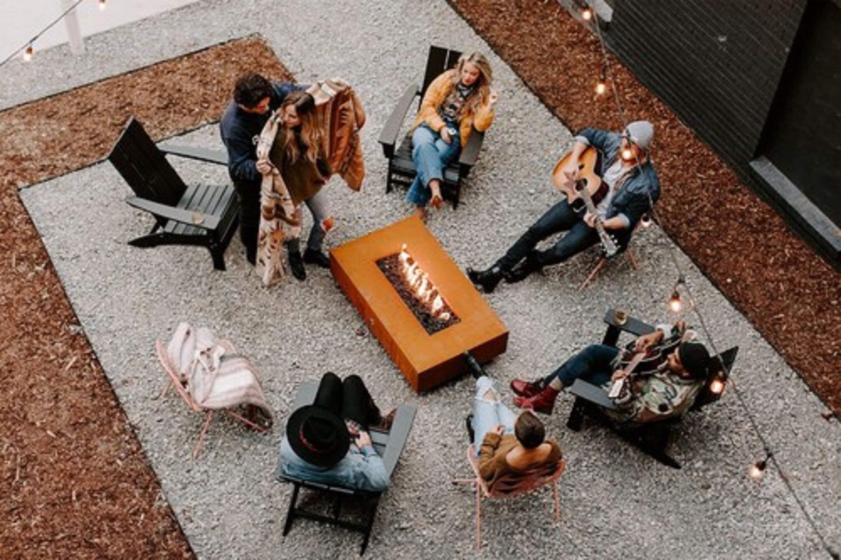 Een groep mensen zit rond een vuurkuil in een openlucht omgeving, met banken en stoelen.