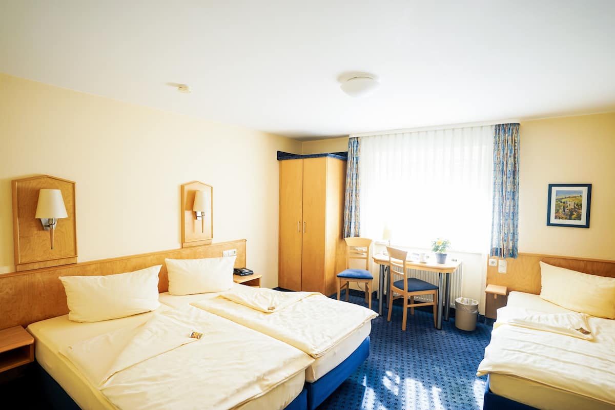 Une chambre d’hôtel avec deux lits, un bureau, une chaise, une lampe, une armoire et une fenêtre avec des rideaux.