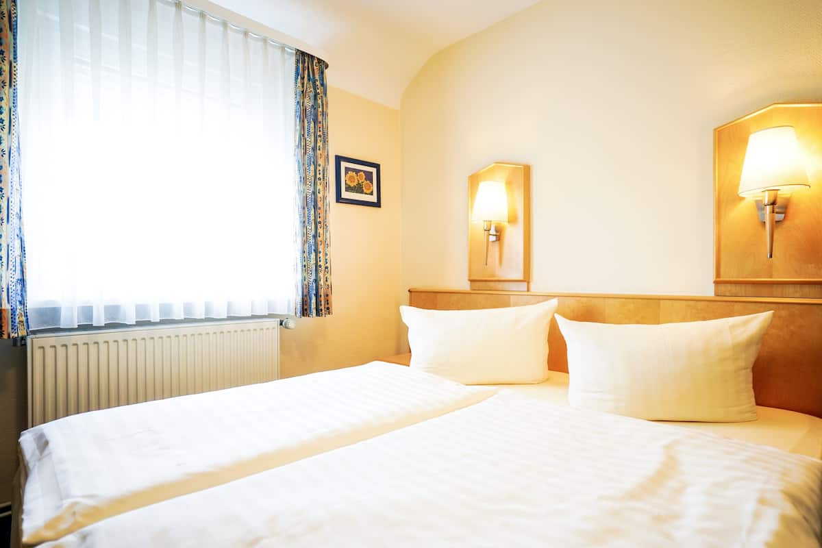 Une chambre d’hôtel avec un grand lit, deux lampes de chevet, une fenêtre avec des rideaux légers et un radiateur.