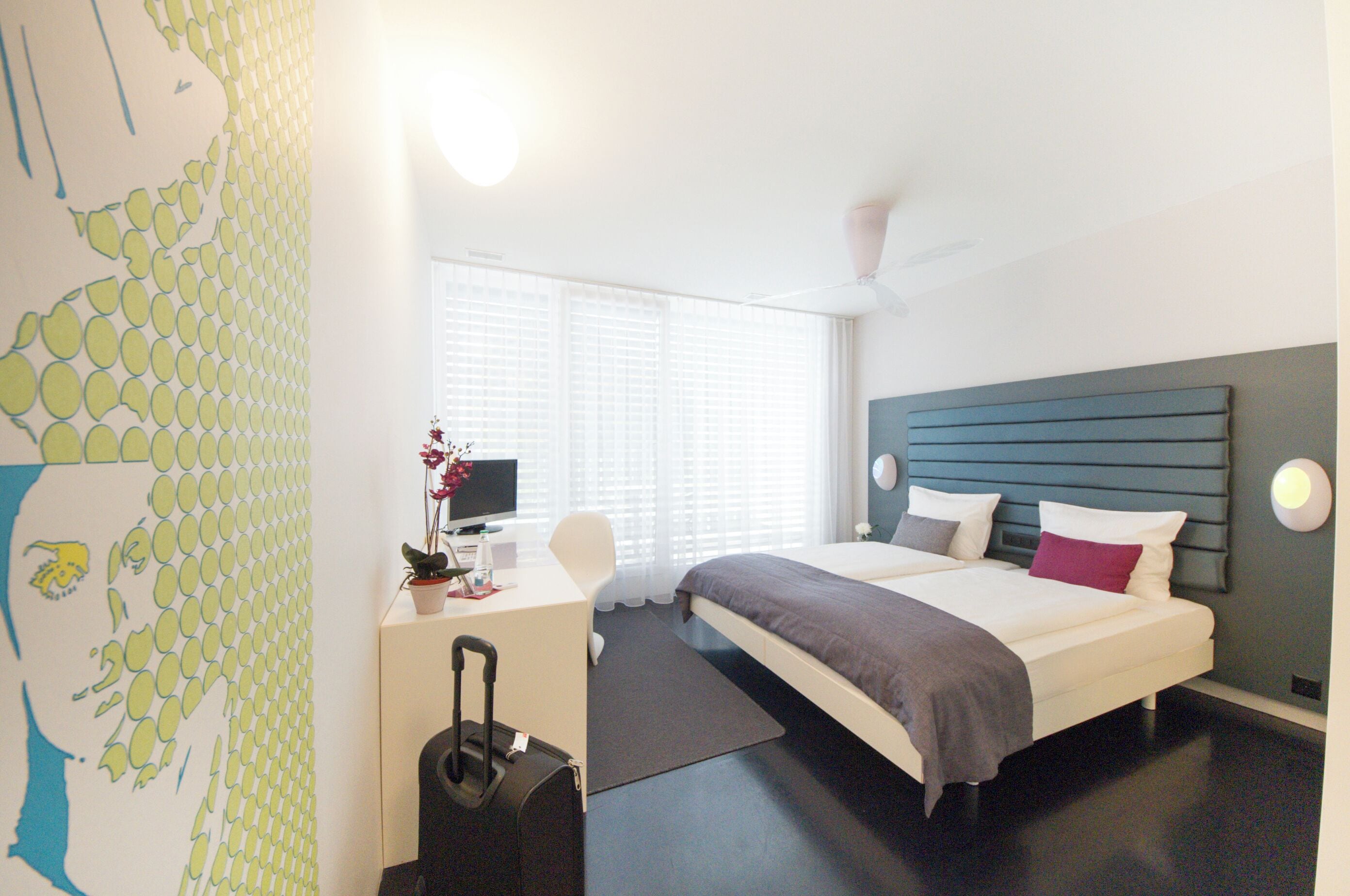 Habitación de hotel con cama, escritorio, silla, maleta y ventana con persianas.
