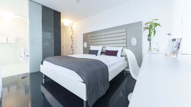 A modern bedroom with a large bed, a bedside table, a wall-mounted TV, and a potted plant. - Sky Motel (Oberriet)