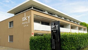 A modern building with a flat roof and a sign that reads 'SKY MOTEL'. There are windows with blinds, a fire hydrant, and a hedge in front.
