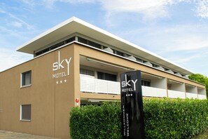 Un edificio moderno con techo plano y un letrero que dice 'SKY MOTEL'. Se ven ventanas con persianas, un hidrante y un seto delantero.