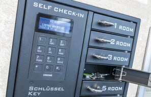 A self-check-in kiosk with a keypad and a digital display. It has numbered keys for room selection and a metal handle for opening a drawer. - Sky Motel (Oberriet)