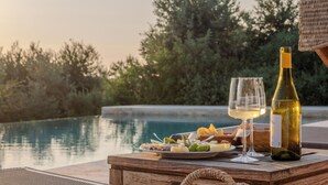 A wooden table with a bottle of wine, two glasses, and a plate of food by a poolside. - Podere Le Mezzelune (Bibbona)