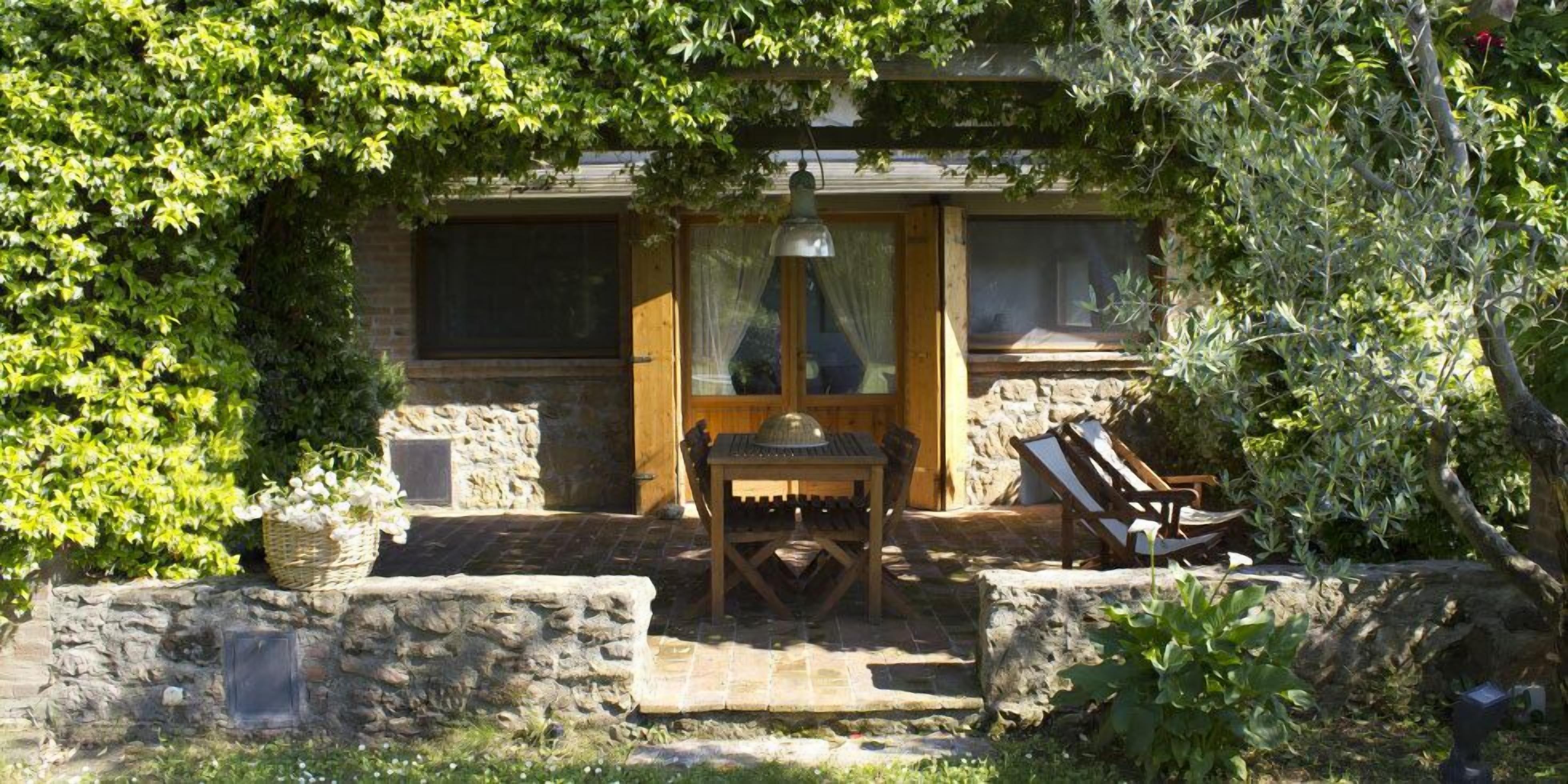 Un bâtiment en pierre avec une porte et des fenêtres en bois, une table avec des chaises et un mur en pierre orné d’un panier de fleurs.
