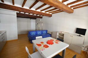 A room with a wooden ceiling, a white table with a floral tablecloth, a blue sofa, a white cabinet, and a television on a stand. - Egidio studio apartment 2 (Florence)