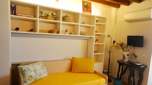 Een kamer met een witte slaapbank voorzien van een gele hoes, een boekenplank met diverse spullen, een bijzettafeltje met een vaas en een aan de muur bevestigde televisie.