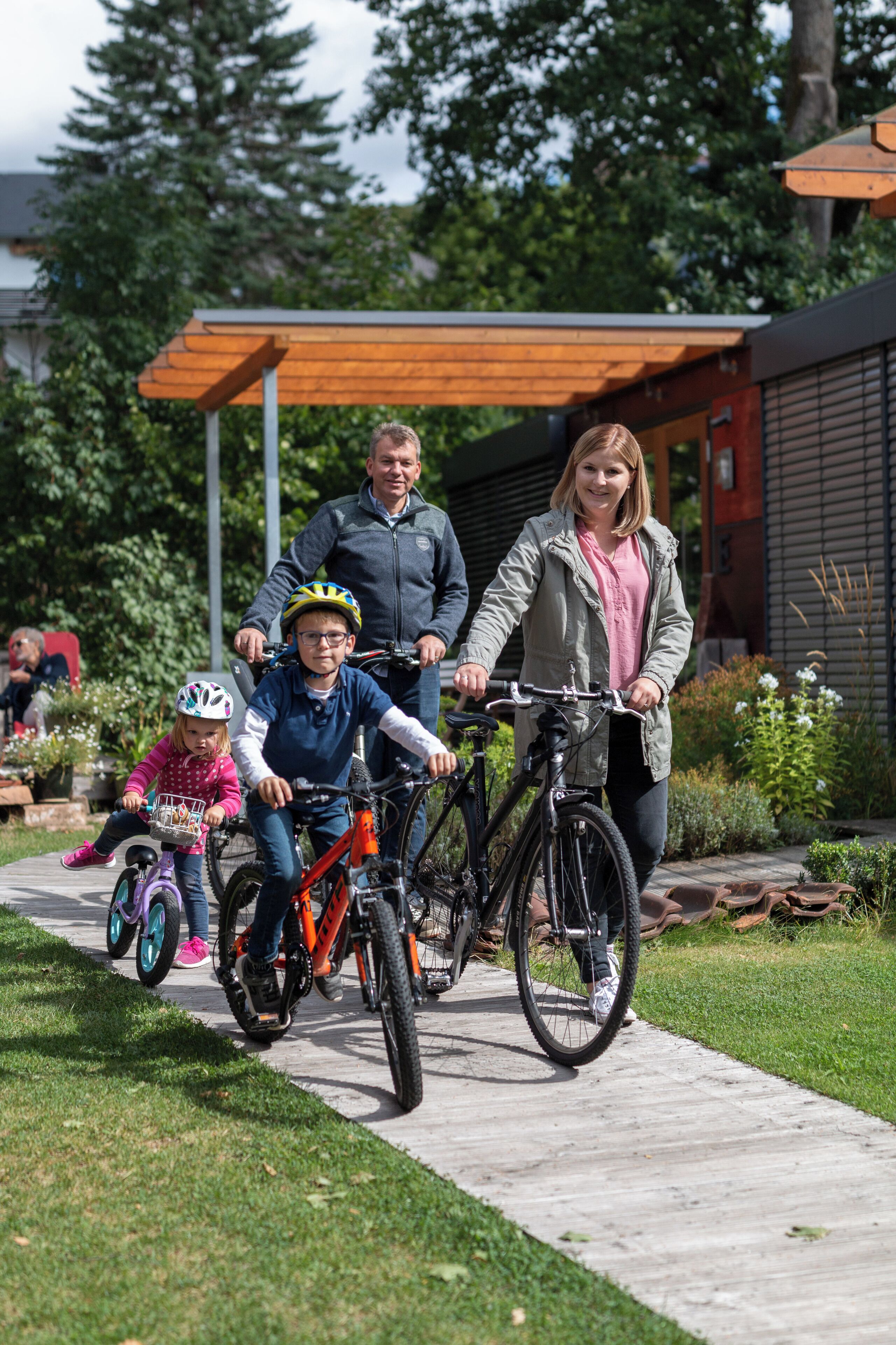 A family with bicycles on a paved path, a wooden pergola, and a building with a sloped roof in the background.