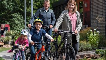A family with bicycles on a paved path, a wooden pergola, and a building with a sloped roof in the background.